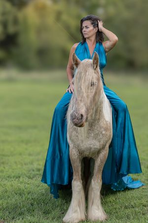 A graceful long-haired miniature horse poses in a grassy field with someone wearing a flowing teal dress.