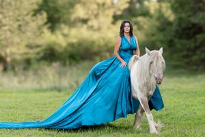 A flowing turquoise ball gown trails elegantly behind a white horse in a sunlit meadow.