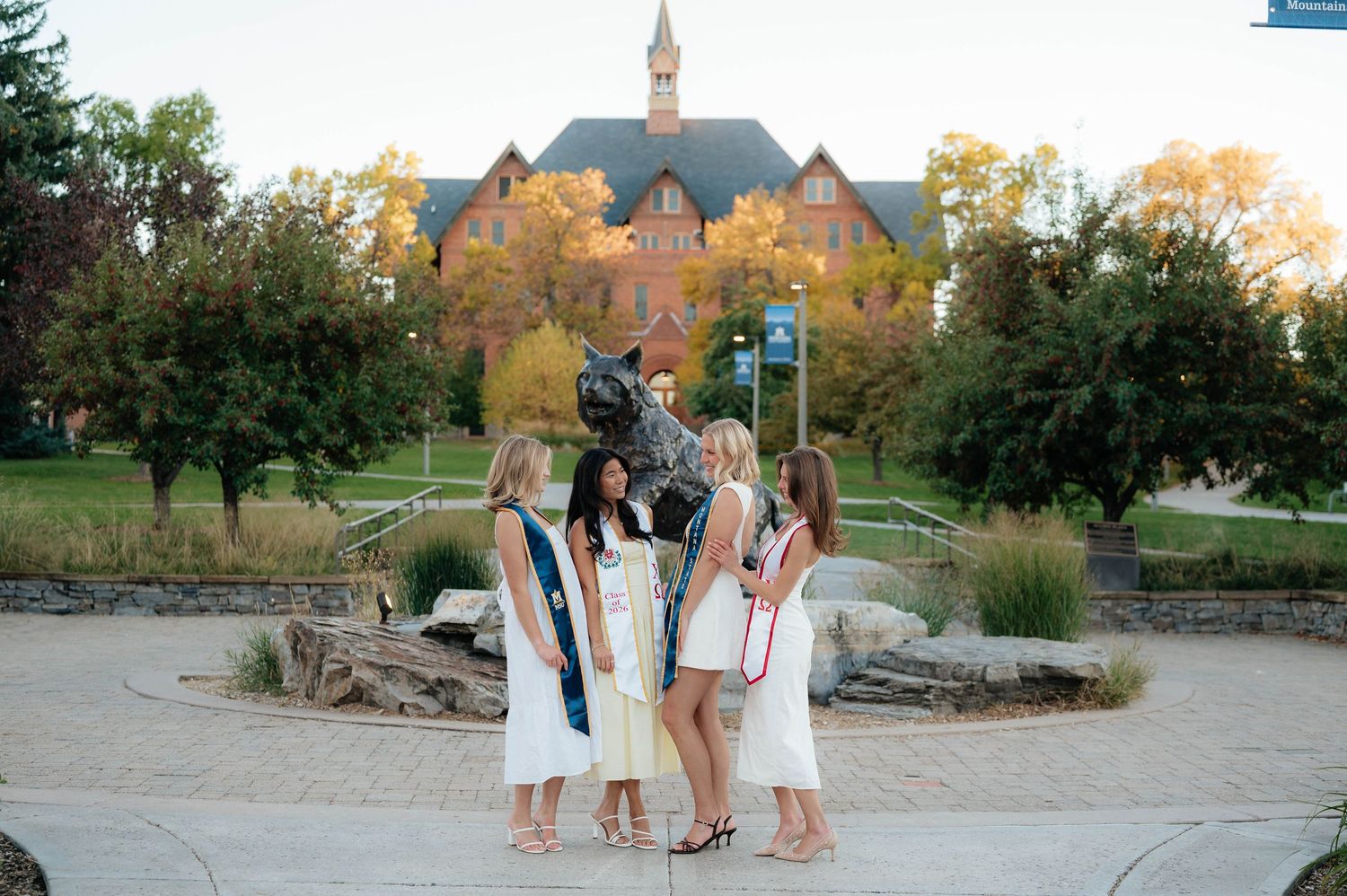 Group of women in white dresses with graduation sashes posing near campus bobcat statue at sunset.