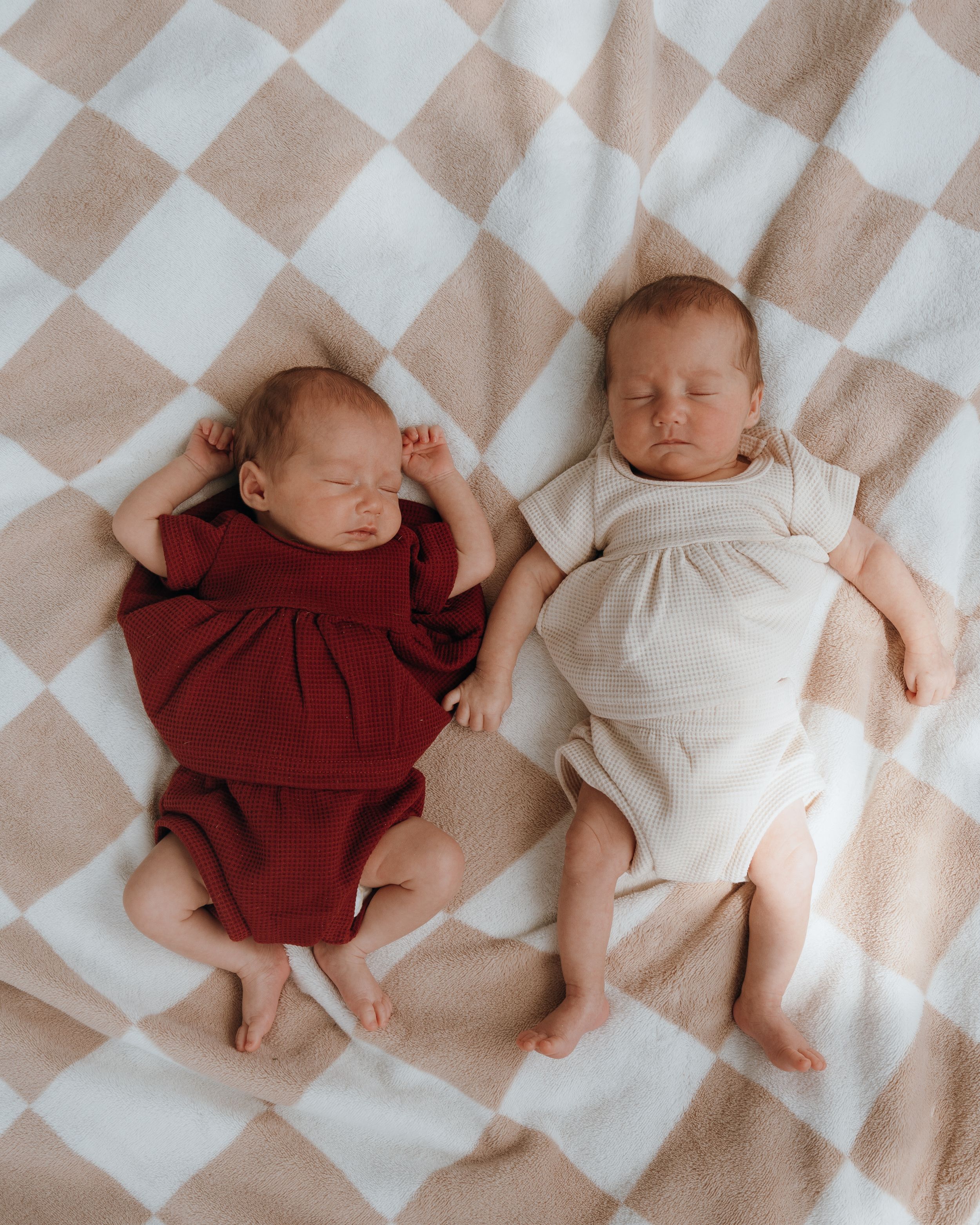Two infants peacefully sleeping side by side on a checkered blanket in burgundy and cream colored outfits.