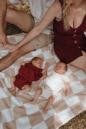 Two babies napping on a picnic blanket with adults sitting nearby on a sunny day.