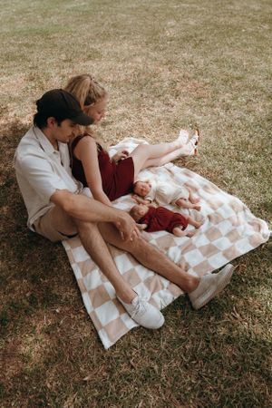 A couple lounging on a checkered blanket outdoors with two sleeping babies.