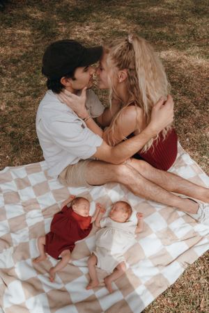 A couple sharing a romantic moment on a picnic blanket while two babies sleep peacefully below.