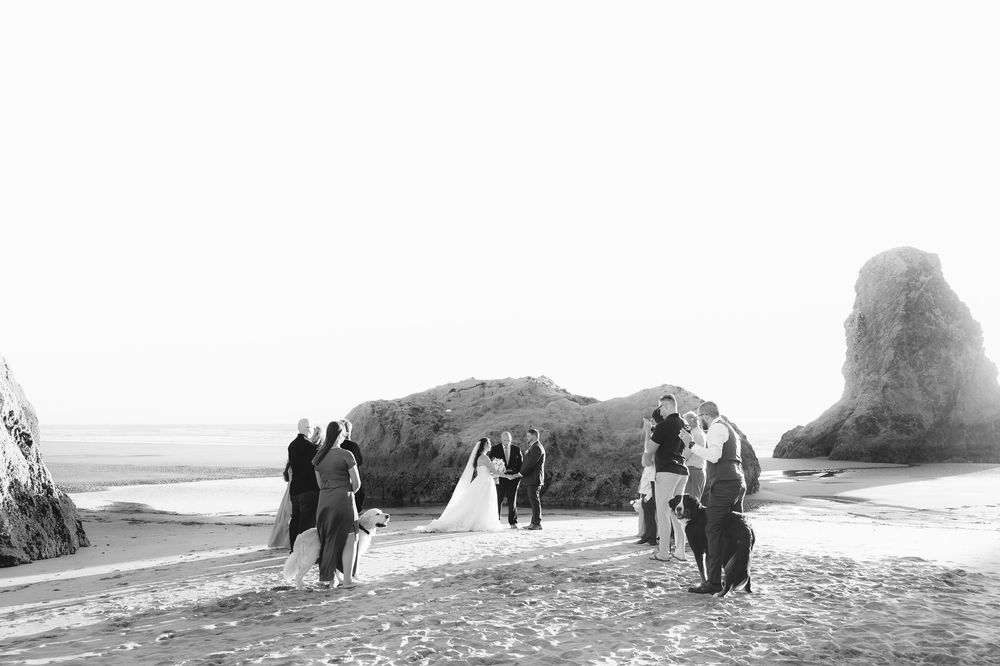 Black and white beach wedding ceremony with couple, guests, and dramatic rock formation by the ocean.
