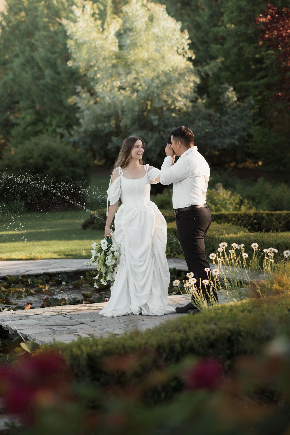 Bride and groom share a romantic moment in a lush garden, bride holding white flowers in an elegant gown.