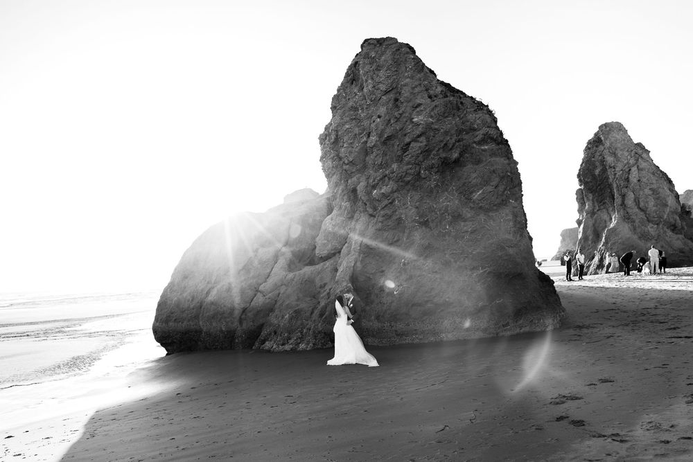 Black and white beach photo with dramatic rock formations, sun flare, and a bride in a white dress on sandy shore.