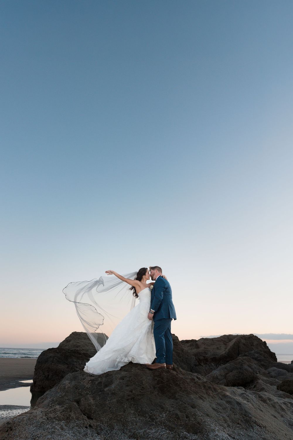 Bride and groom kissing on coastal rocks at sunset, bridal veil flowing in the wind against a clear blue sky.