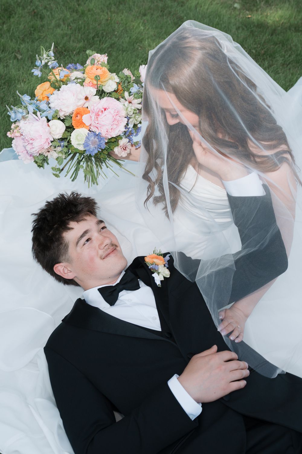 Bride and groom lying on grass, sharing a romantic moment under a white veil, with a colorful floral bouquet nearby.