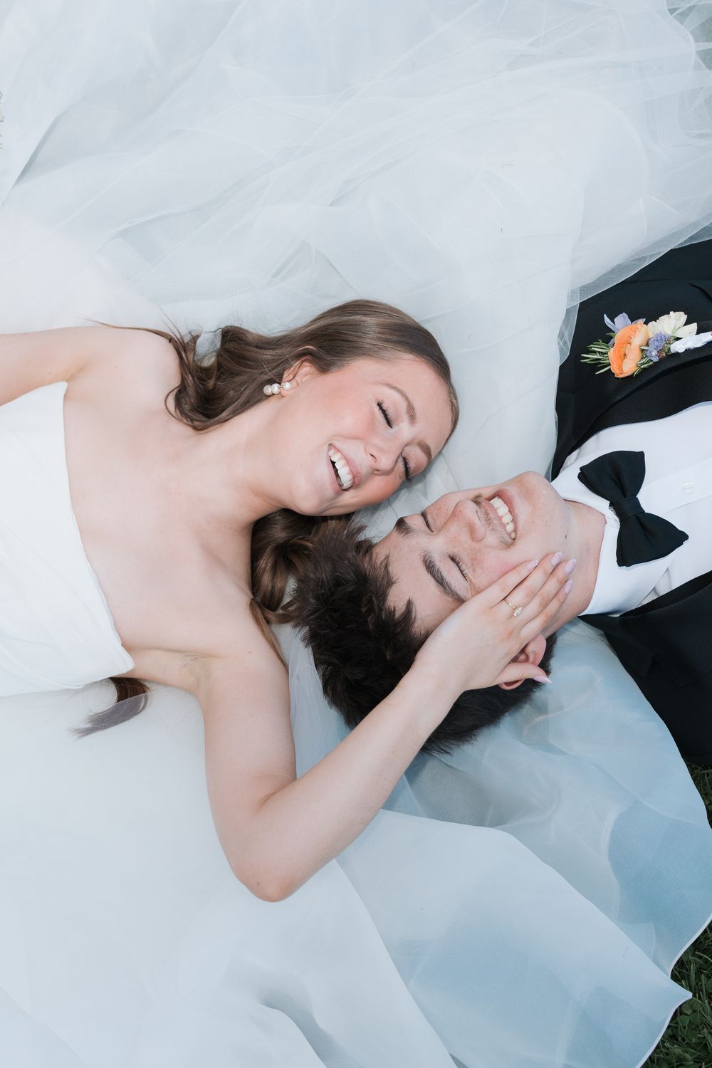 Joyful bride and groom in wedding attire lying on grass, laughing together in a romantic top-down portrait.