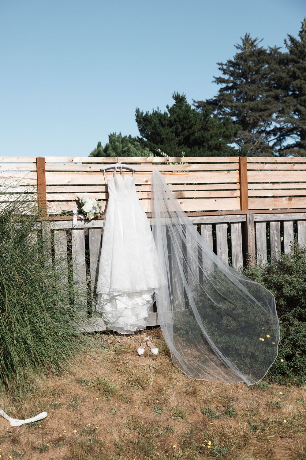 White wedding dress with long veil hanging on wooden fence outdoors, with bridal shoes below on a sunny day.