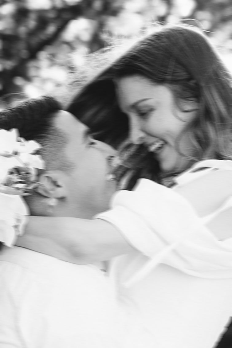 Black and white photo of a smiling couple embracing outdoors, surrounded by blossoming trees.