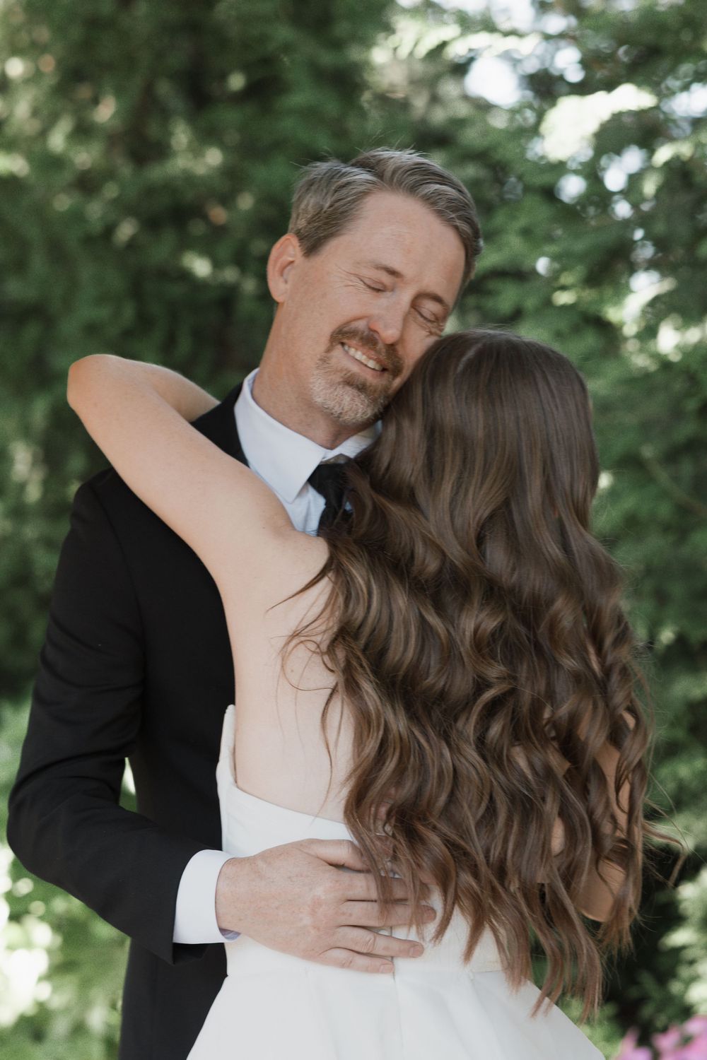 A groom in a black tuxedo embraces his bride in a white gown, sharing a tender moment outdoors.