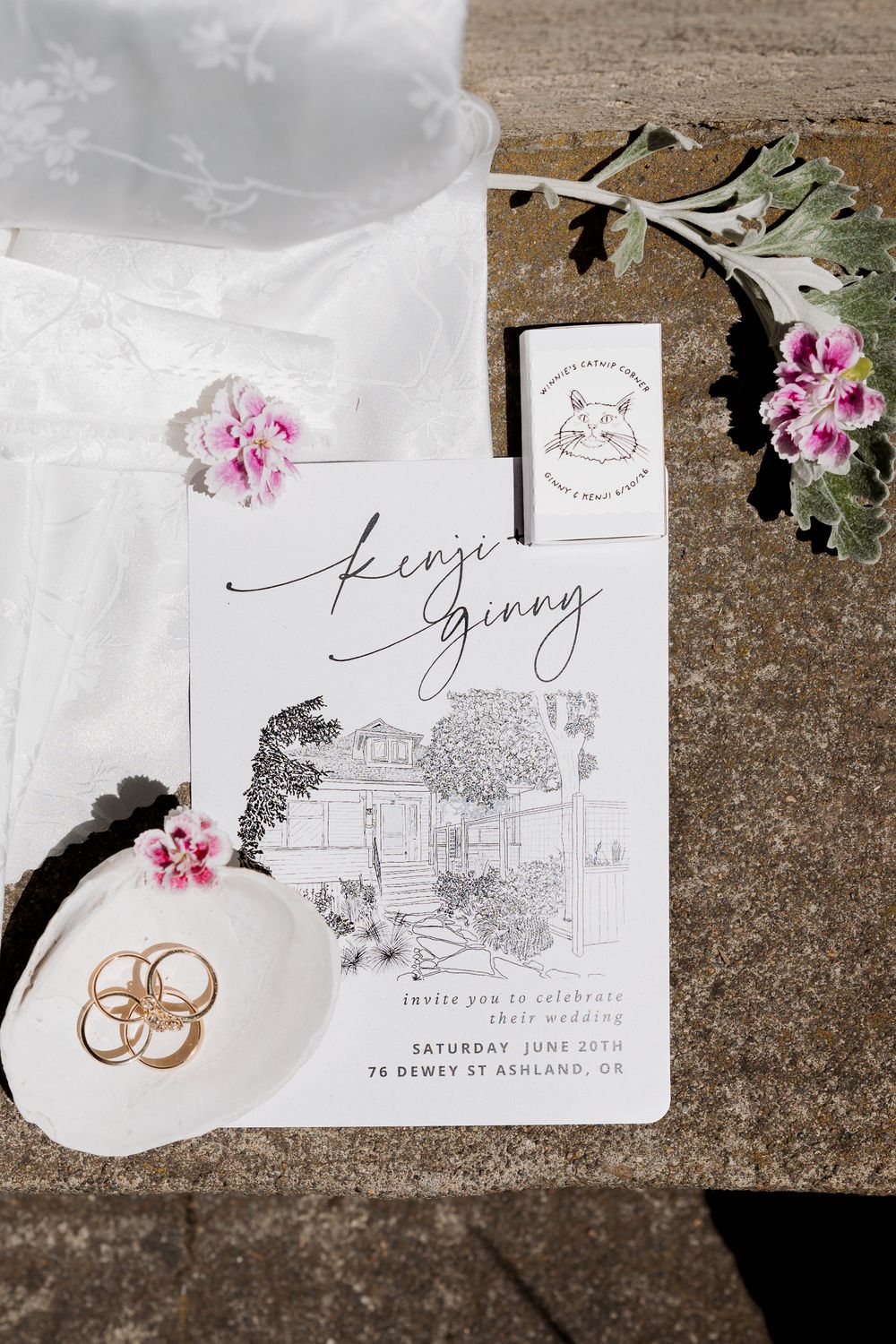 Elegant wedding invitation for Kenji and Ginny with hand-drawn venue sketch, gold rings, pink flowers, and matchbook on stone surface.