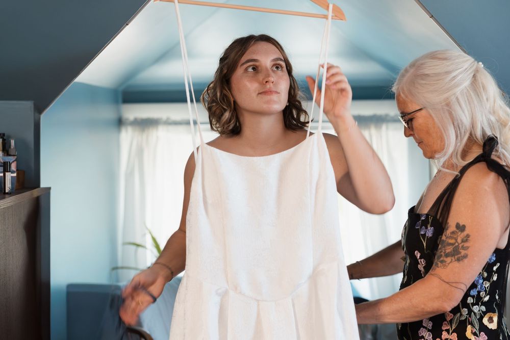 Older woman helping young bride try on white wedding dress in a blue-ceiling room.