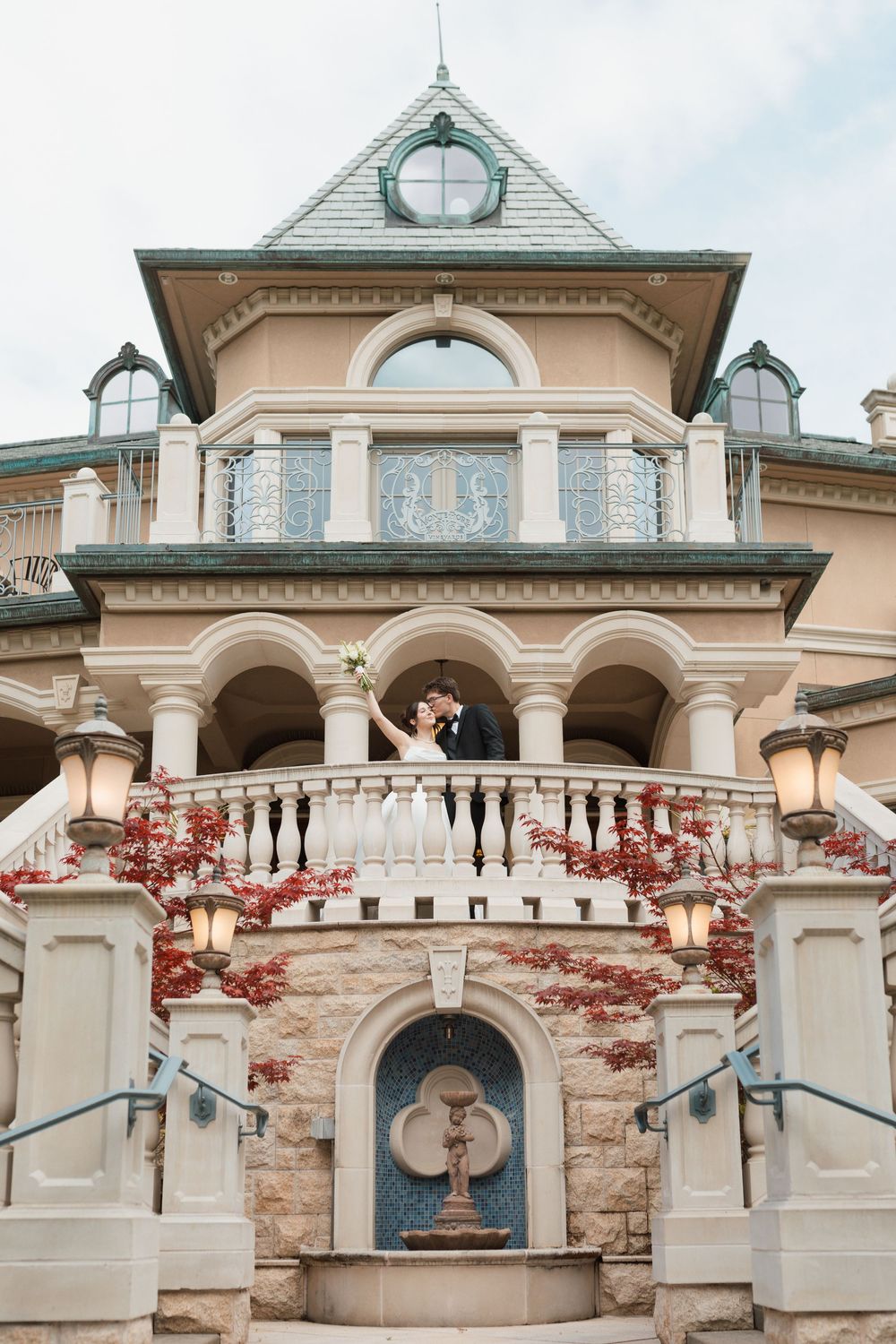 Bride and groom kissing on ornate mansion balcony adorned with red ivy and elegant stone architecture.