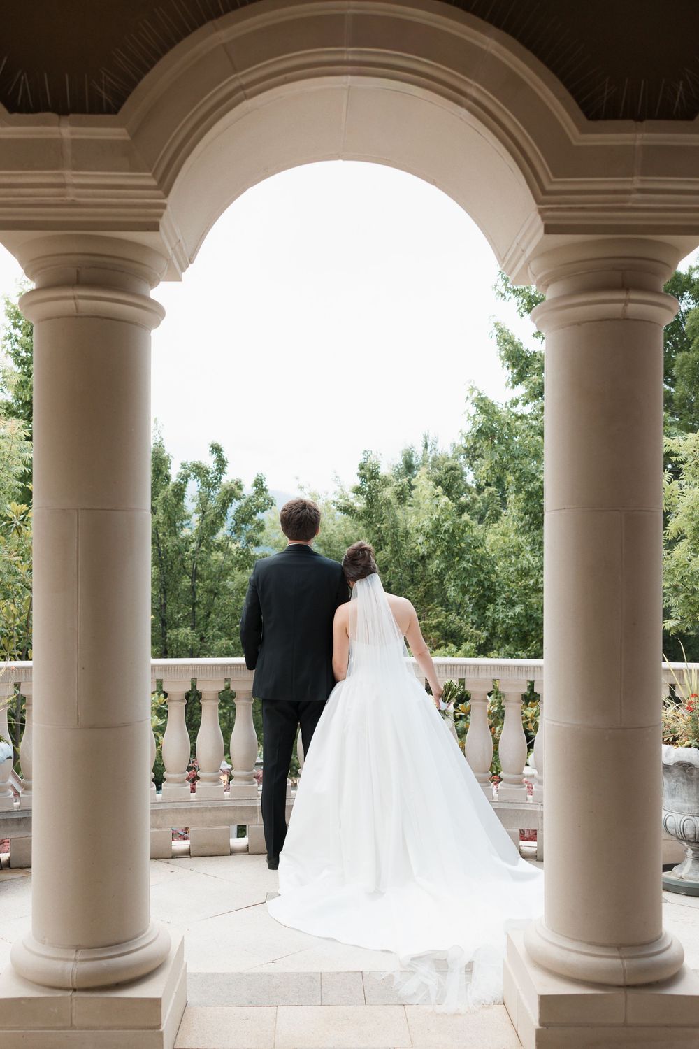 Bride and groom stand between elegant columns on a wedding venue terrace, overlooking lush green gardens.