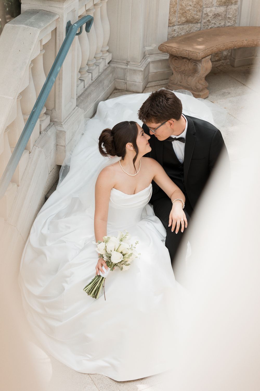Bride and groom sharing a kiss on marble steps, bride holding a white floral bouquet in elegant wedding attire.