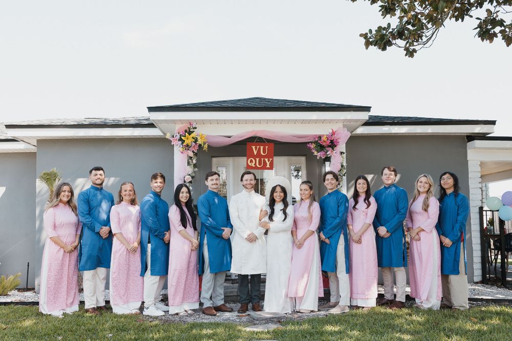 Wedding party in pink ao dai and blue ao dai poses outside a decorated venue with a floral arch.