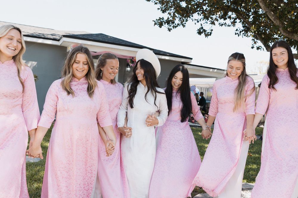 A group of women in matching pink ao dai dresses walk together outdoors, with one woman in white at a wedding.