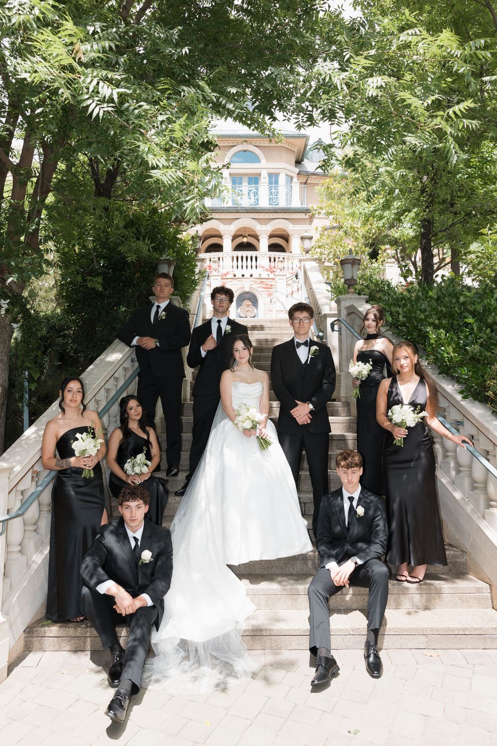 Wedding party poses on elegant stone steps outdoors, bride in white gown with bouquet, attendants in black formal wear.