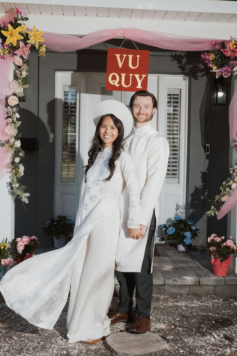Couple in white Vietnamese ao dai traditional attire posing before a Vu Quy sign at a wedding ceremony.