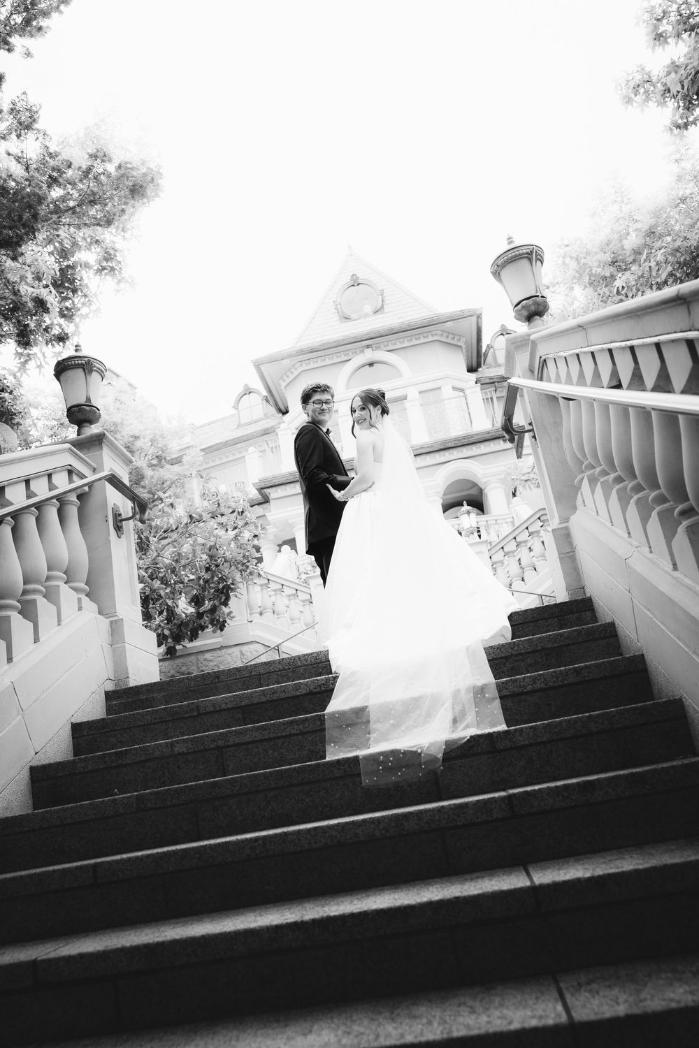 Black and white photo of a bride and groom standing on grand staircase outside elegant mansion.