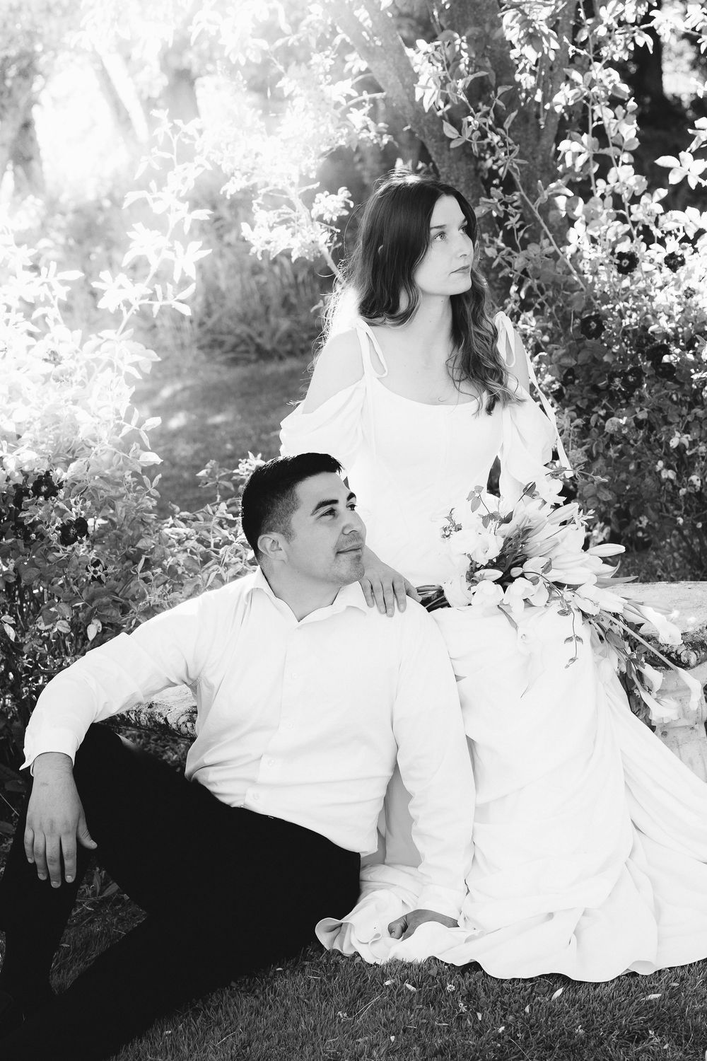 Black and white photo of a bride and groom sitting outdoors under a tree, bride holding a bouquet.