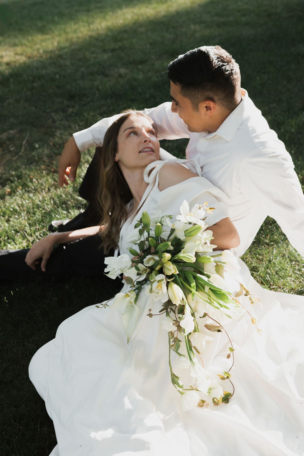Bride in white gown holds floral bouquet while sitting on grass with groom in white shirt during outdoor wedding.