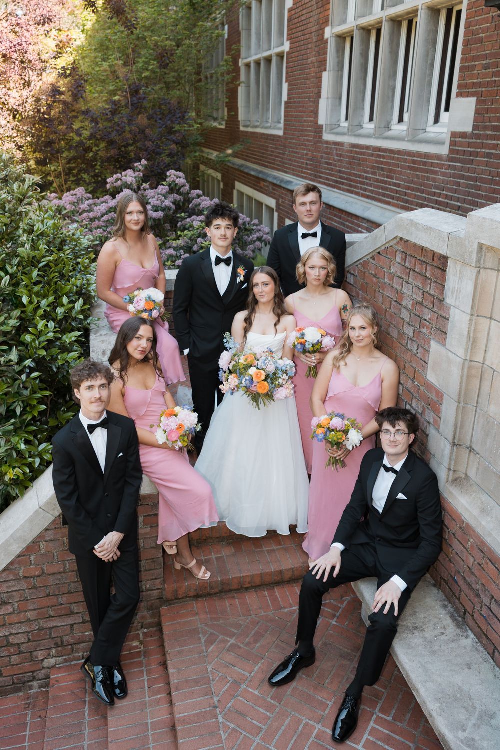 Wedding party poses on brick steps, bridesmaids in pink dresses, groomsmen in tuxedos, bride in white gown holding bouquet.