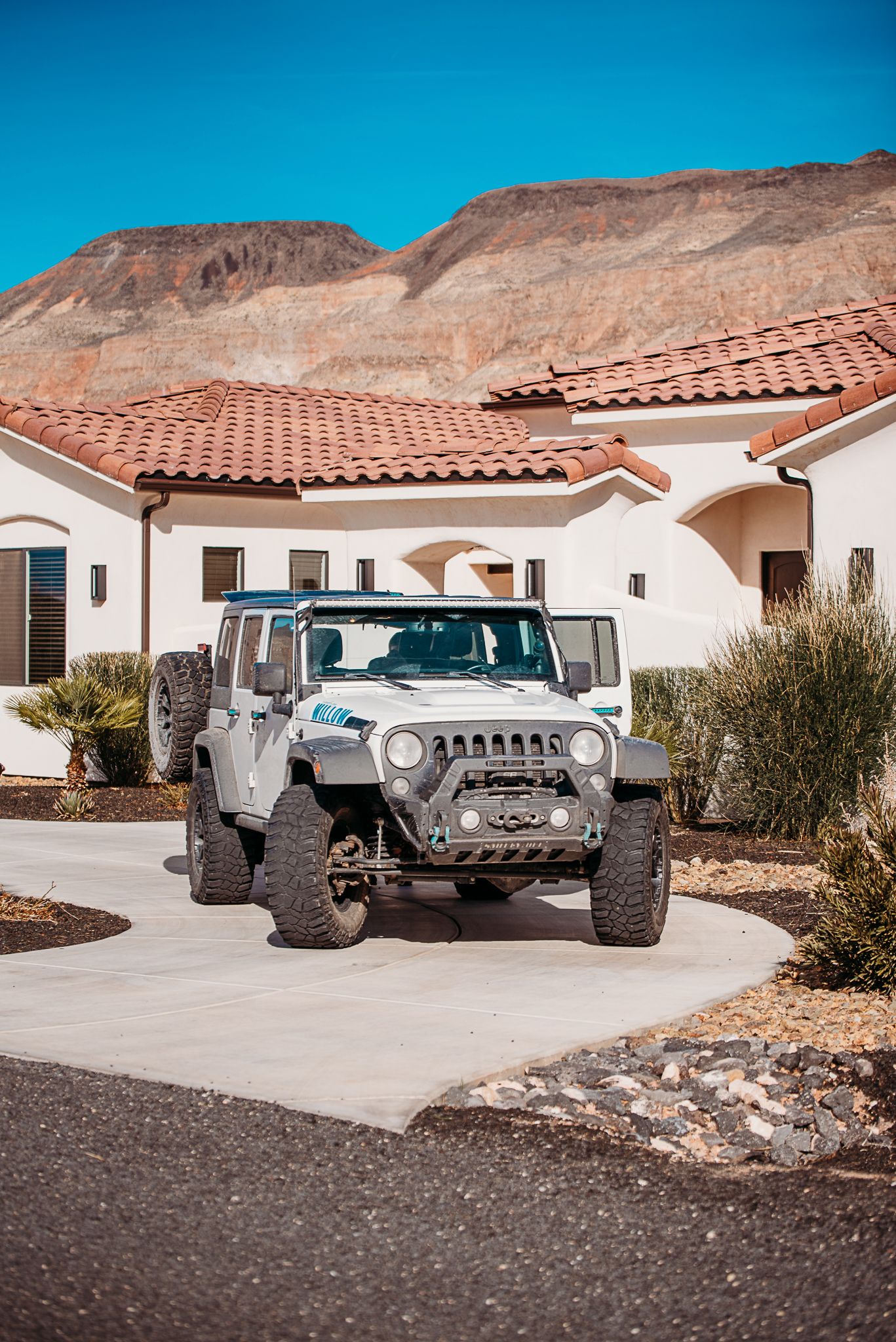 Modified white Jeep Wrangler parked in driveway of Spanish-style house with desert mountains in background.