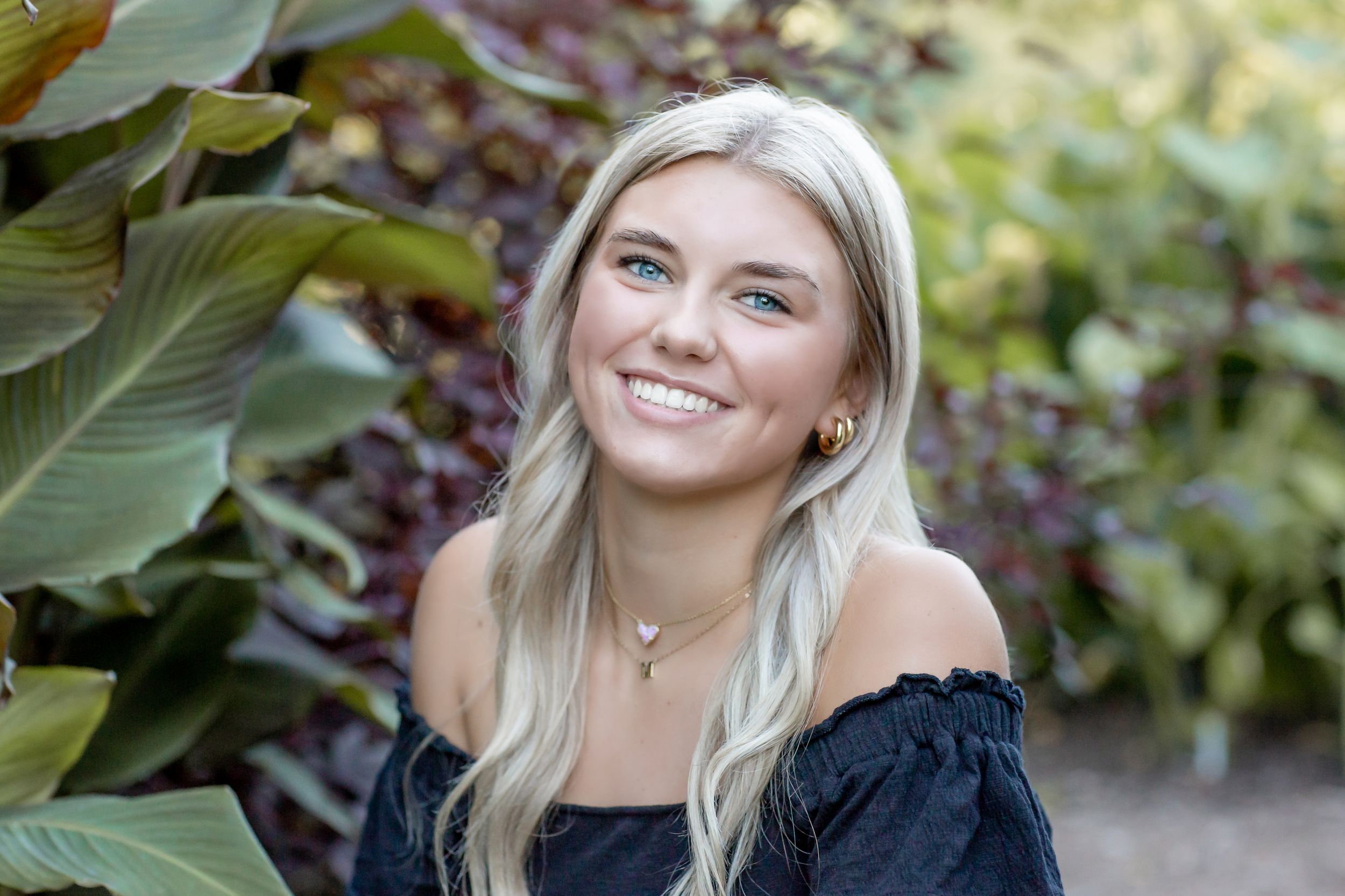 Smiling blonde teen girl in a black off-shoulder top posing near tropical green and purple foliage.