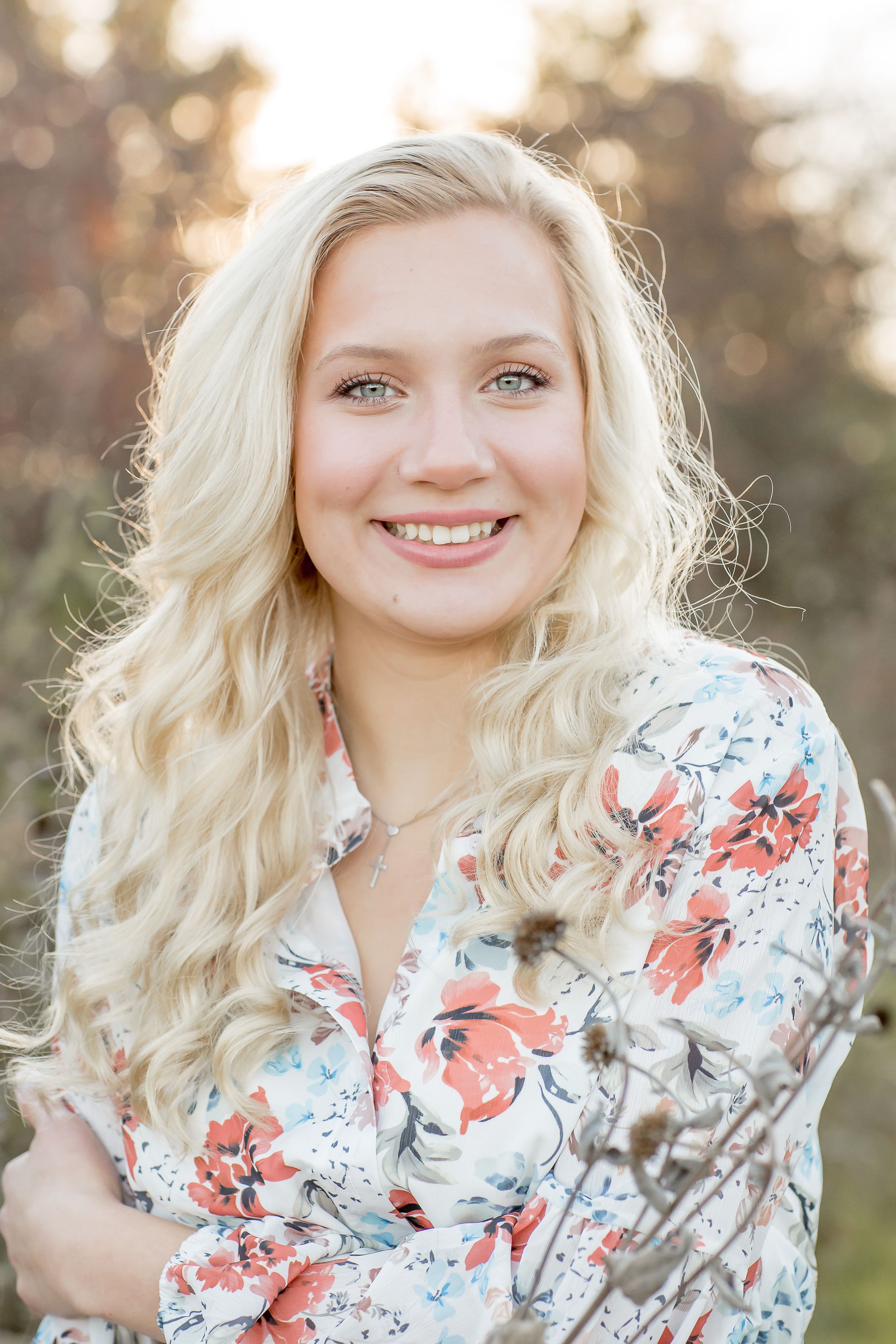 Young blonde woman with long curly hair smiling in floral print blouse outdoors.