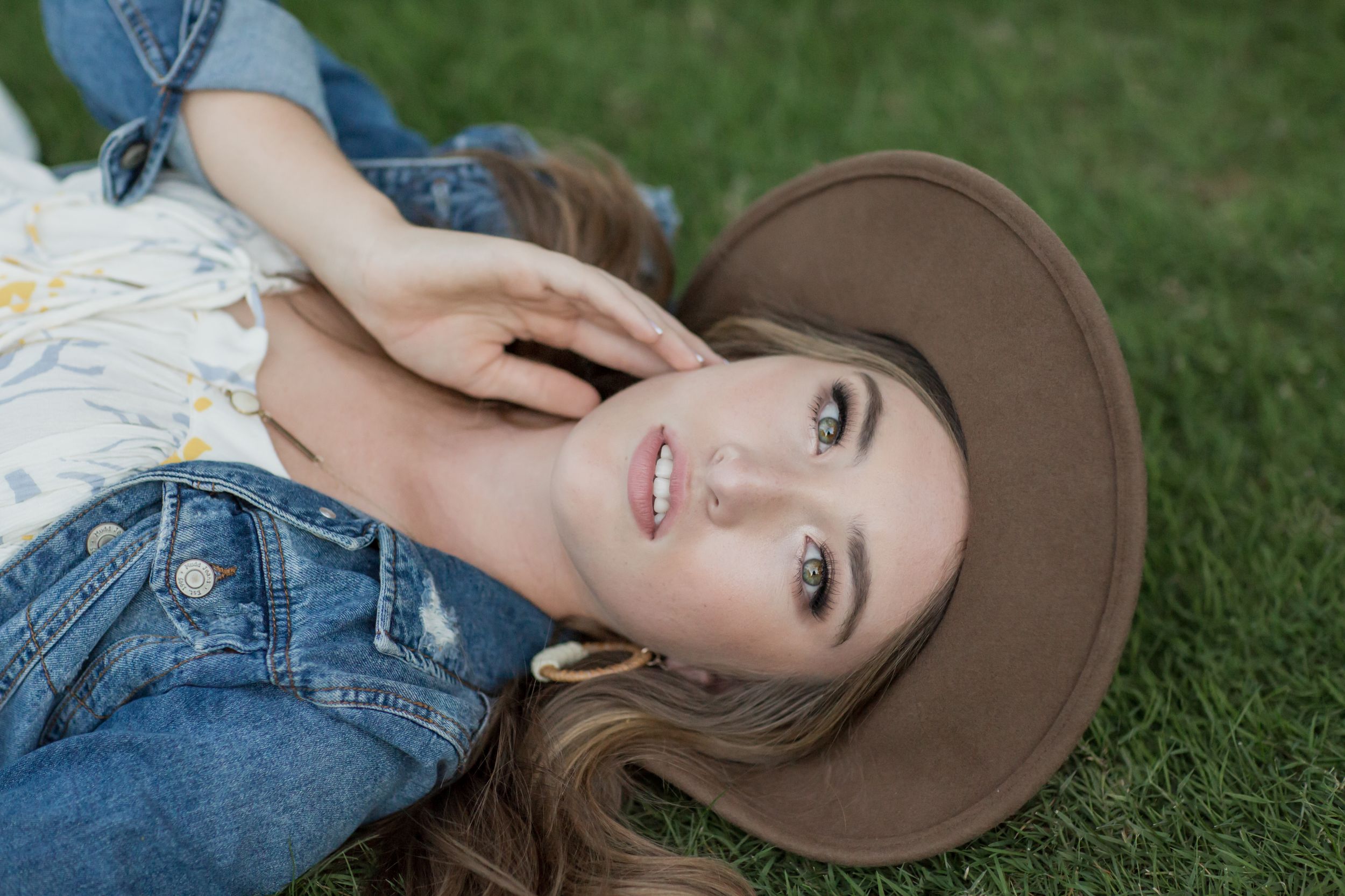 Young woman lying on grass wearing denim jacket and brown hat, looking up at camera.