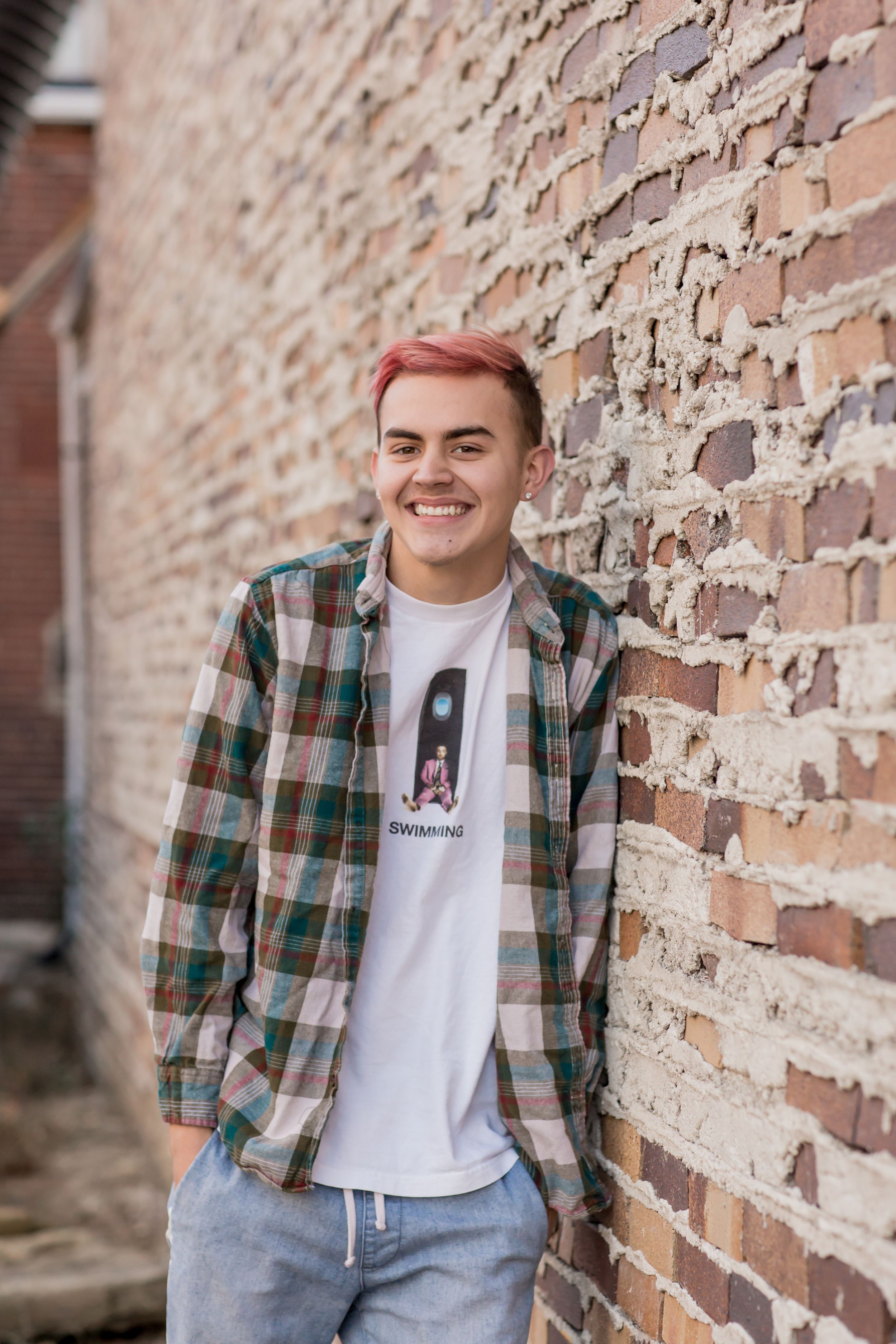 Smiling teen boy with pink hair in a plaid shirt leaning against a rustic brick wall.