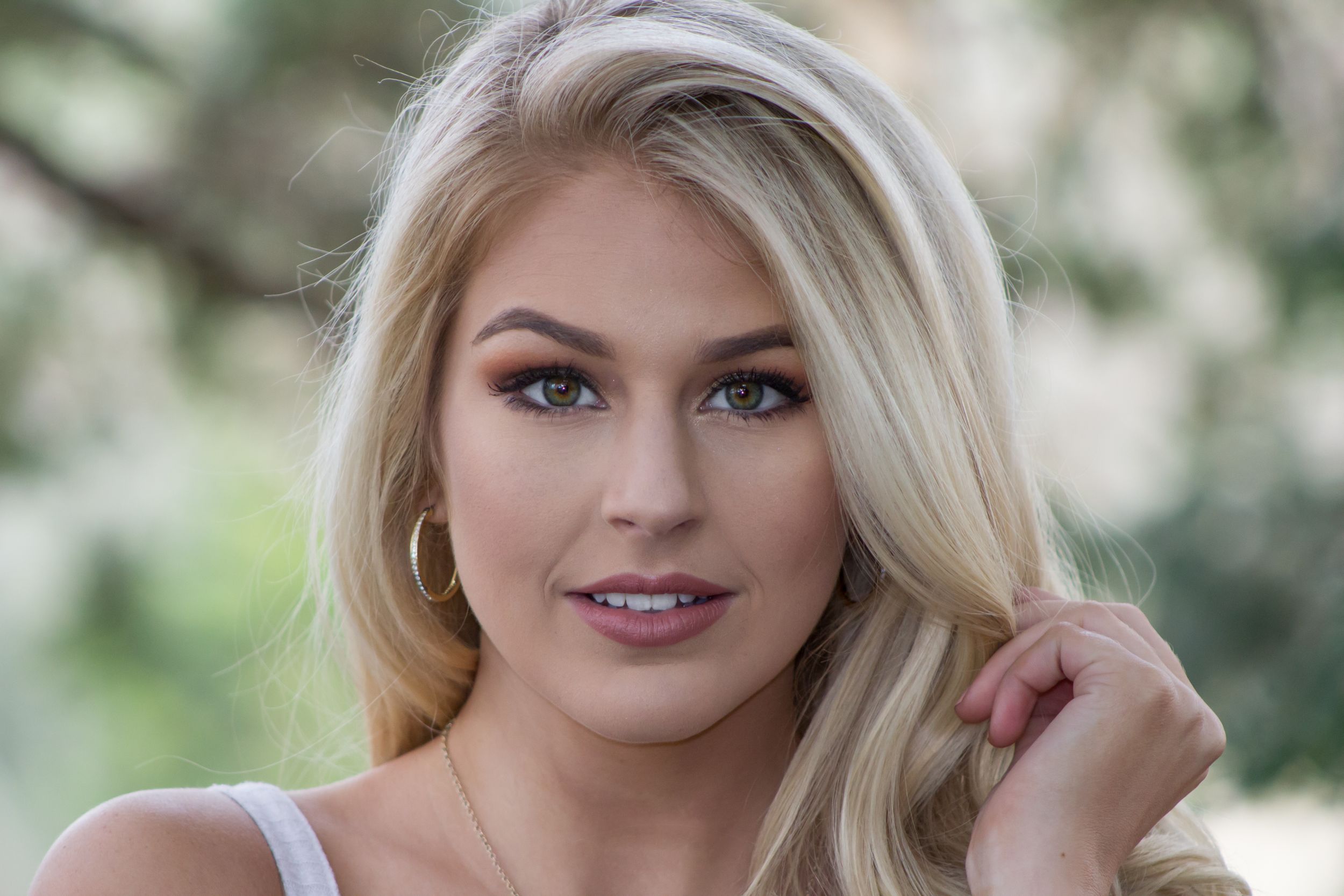 Blonde woman with hoop earrings smiling softly in outdoor bokeh portrait.