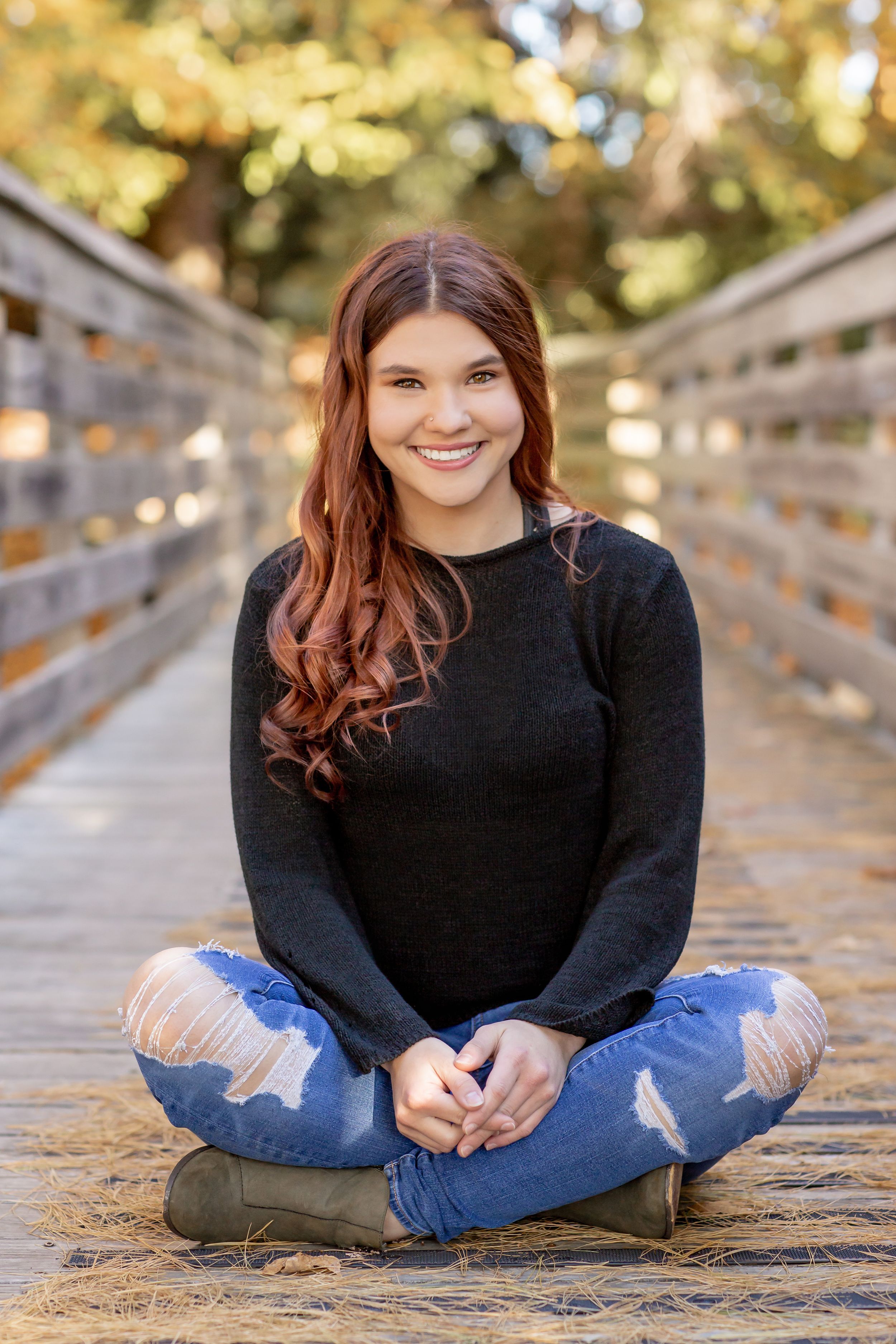 Smiling redhead teen girl in a black sweater and ripped jeans sitting cross-legged on a wooden bridge.