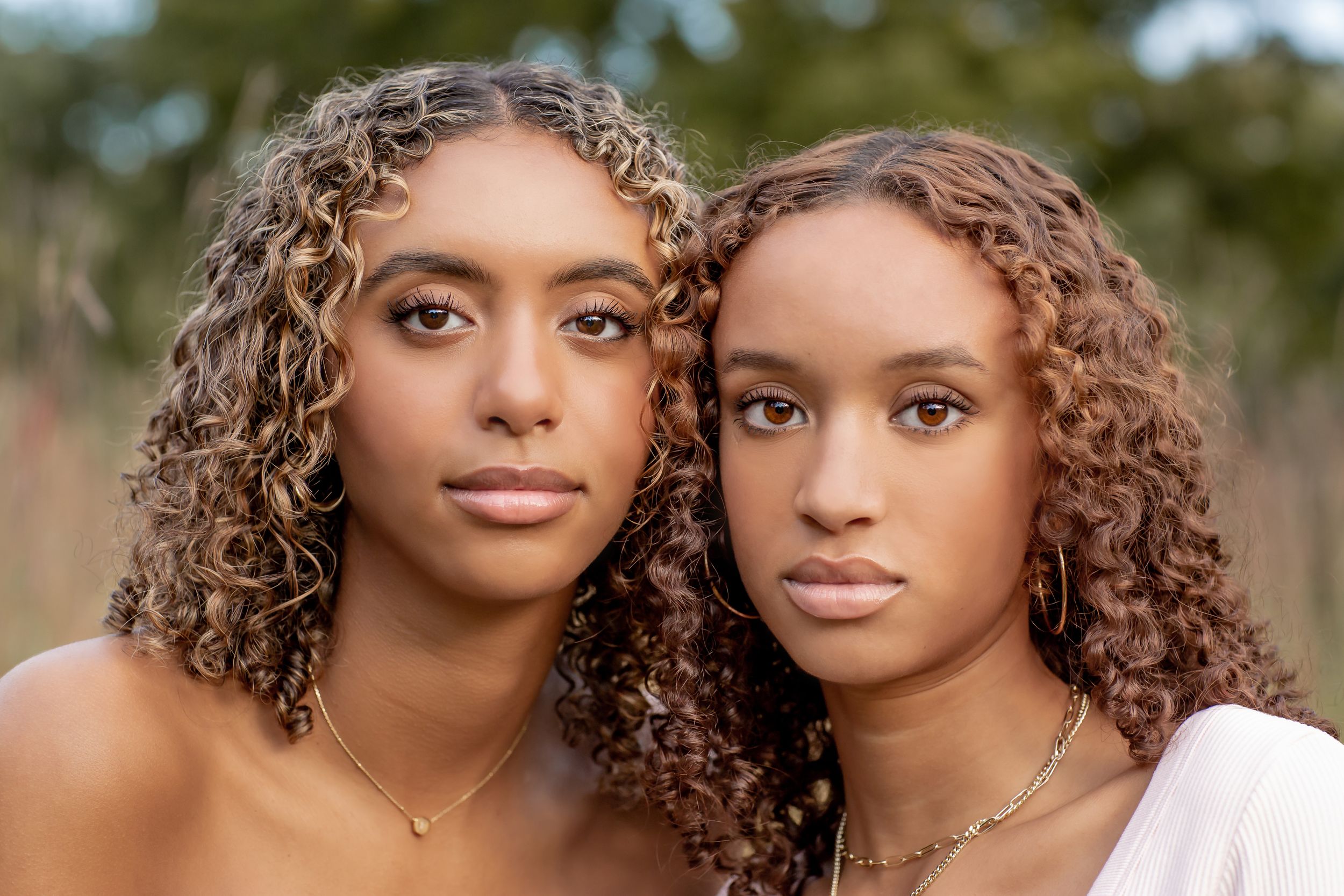 Two young women with curly hair wearing gold jewelry posing closely together outdoors.