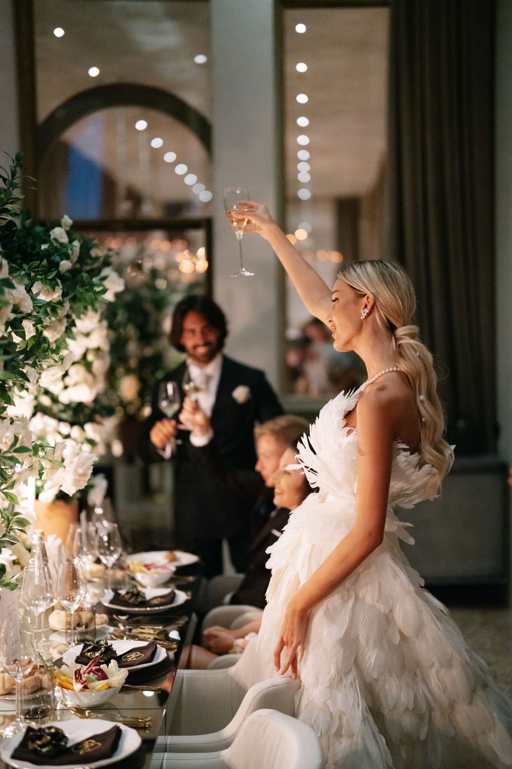 Bride in feathered dress making a celebratory toast at wedding reception.