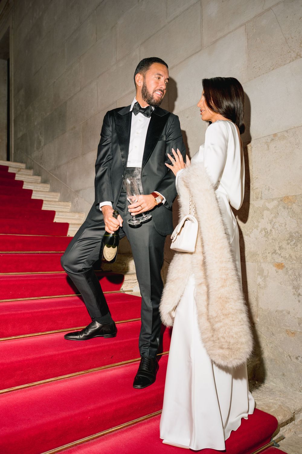 A couple in formal attire share a moment on elegant red-carpeted stairs, with him in a tuxedo and her in a white gown. Venice photographer.