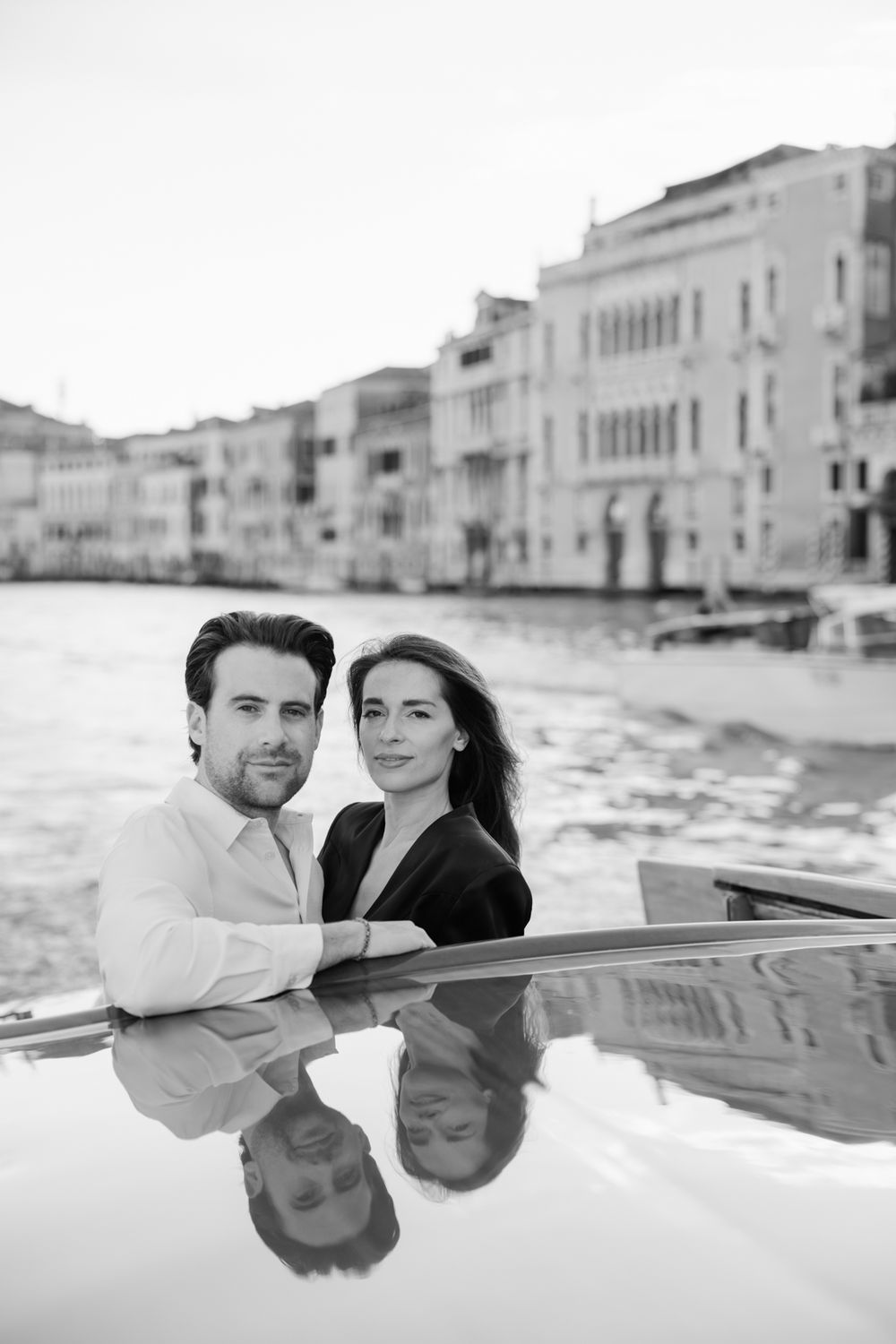 Black and white photo of a romantic couple posing by the Grand Canal in Venice, Italy, with historic buildings reflected in water.