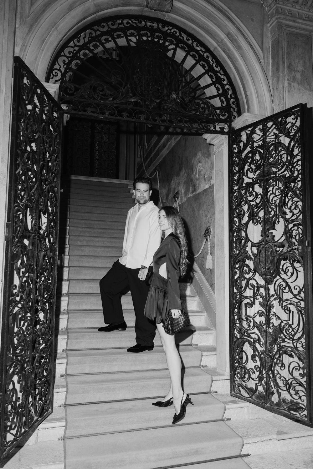 Black and white photo of an elegant couple descending grand staircase through ornate wrought iron doors.