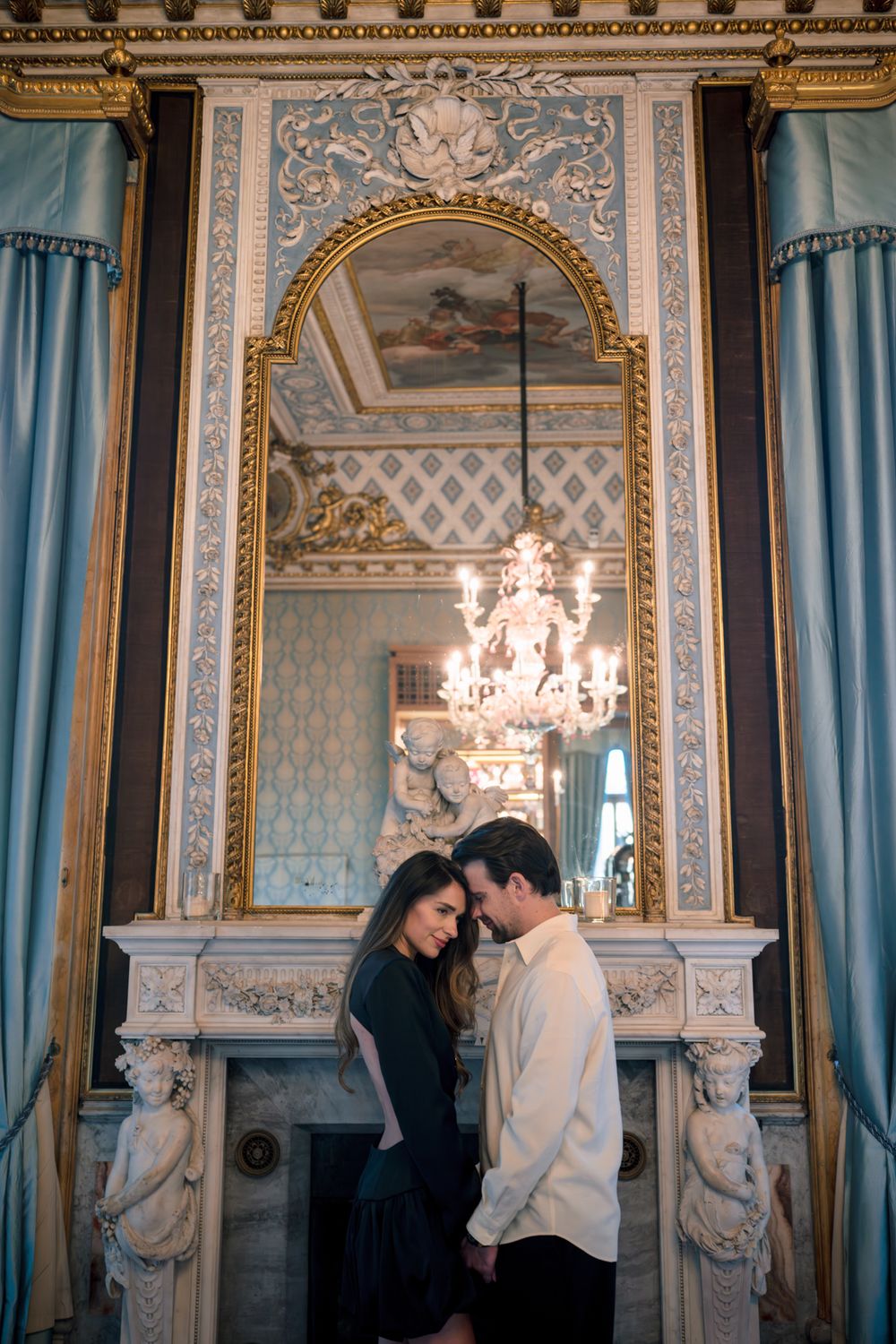 Couple sharing an intimate moment in an ornate room with gold-framed mirror, chandelier, and elegant blue drapes.