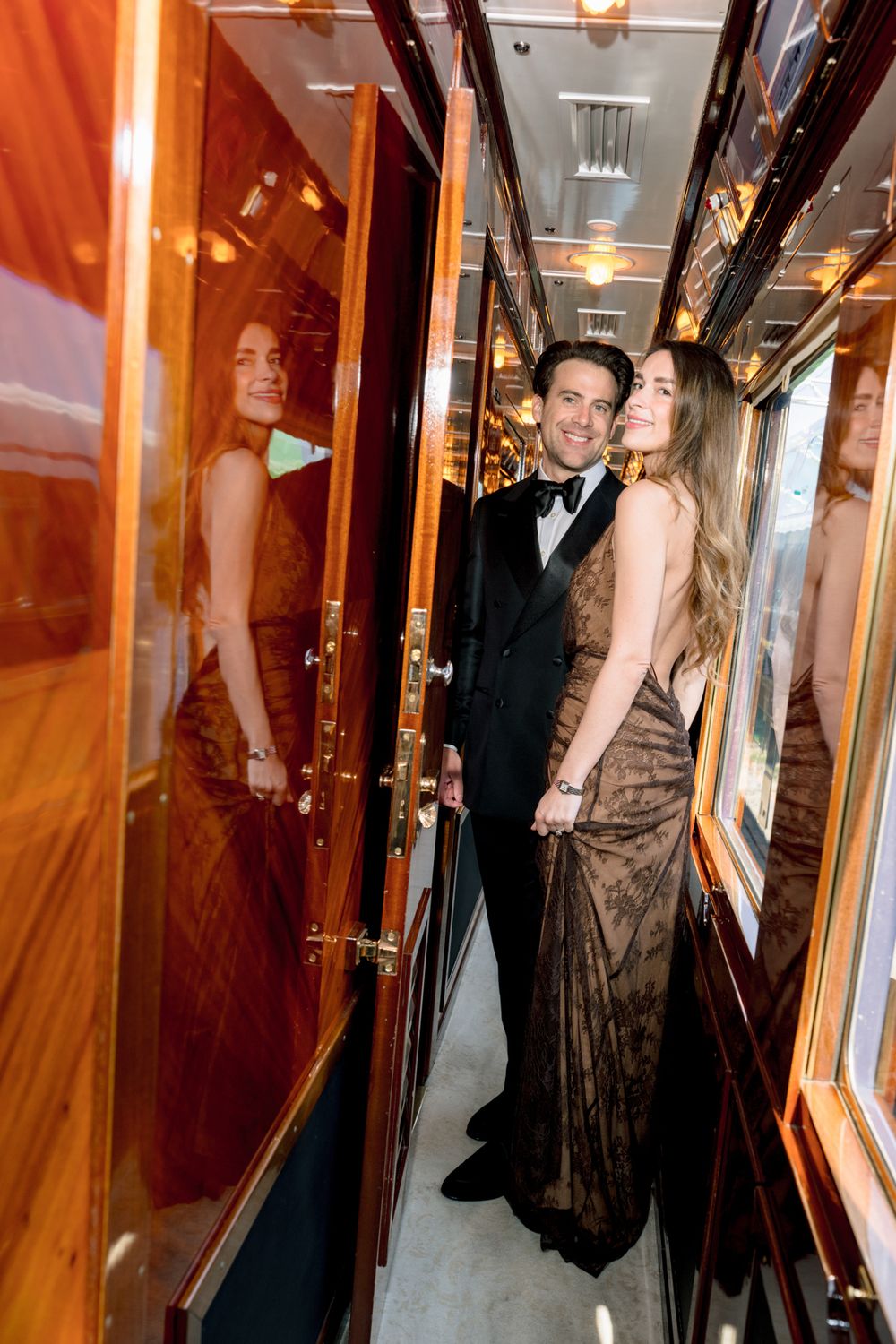 Elegant couple in formal attire posing in a luxurious train corridor with warm wood paneling and mirrored reflections.
