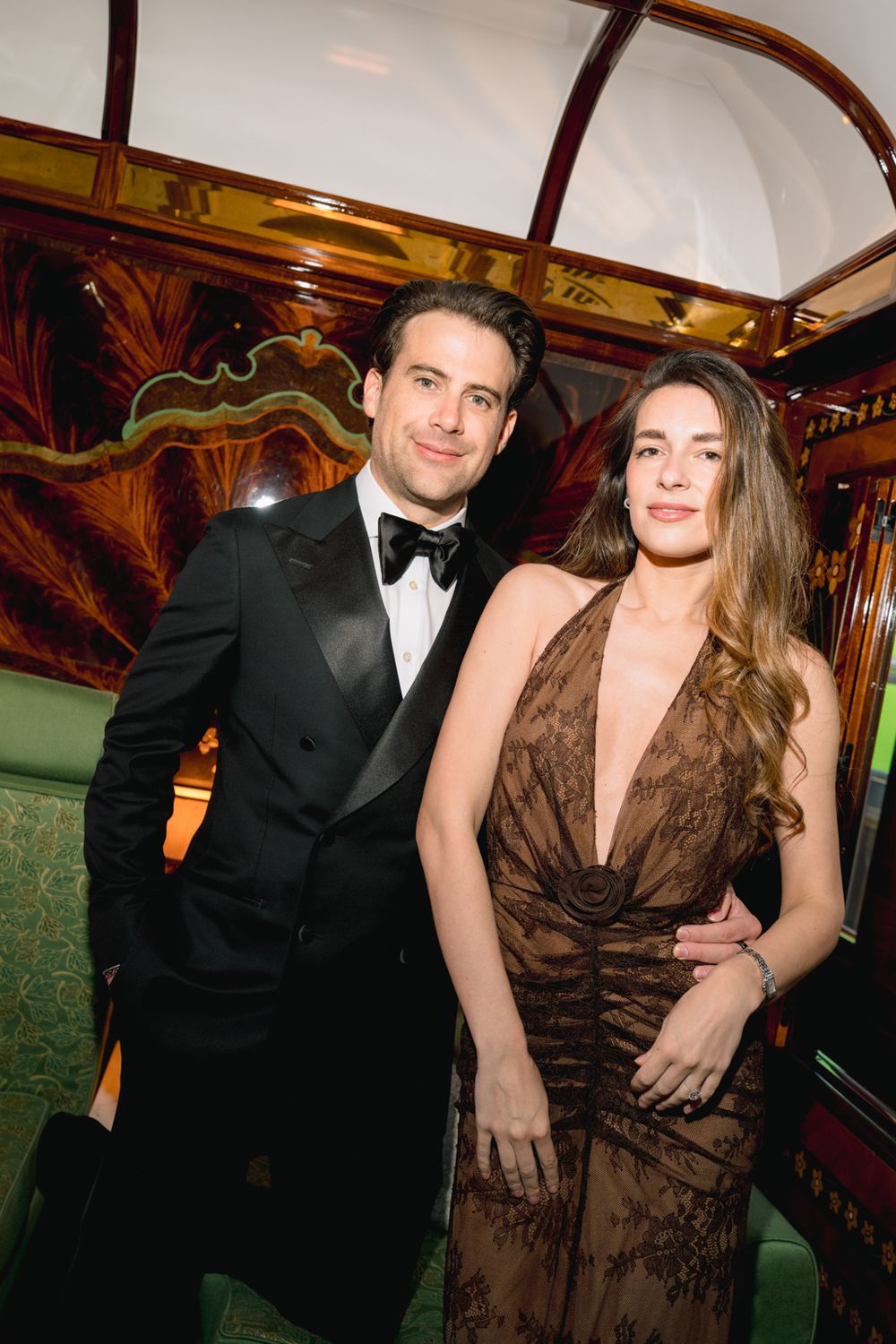 Elegant couple posing at formal event, man in tuxedo and woman in brown lace gown, ornate interior backdrop.