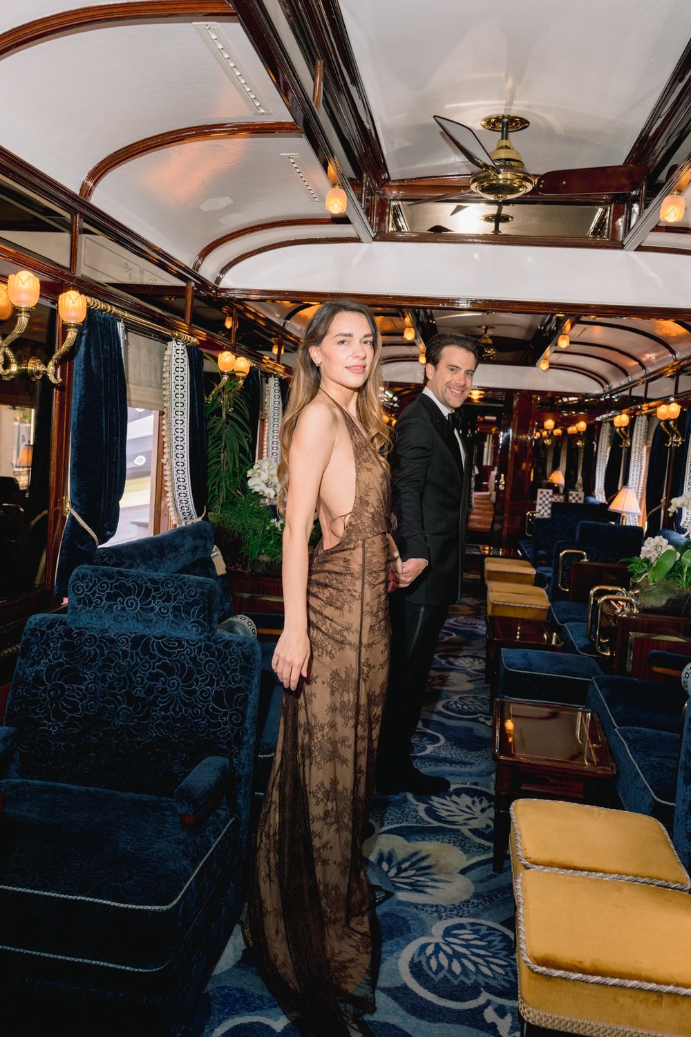 Elegant couple in formal attire posing inside a luxurious vintage train car with blue velvet seating.
