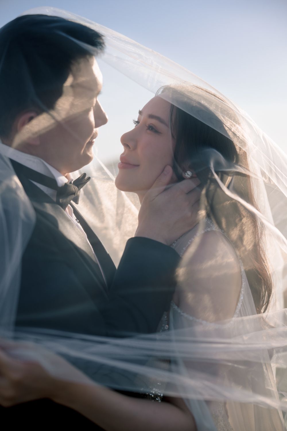 Bride and groom share a romantic moment under a flowing veil, with soft backlight creating an ethereal wedding portrait.