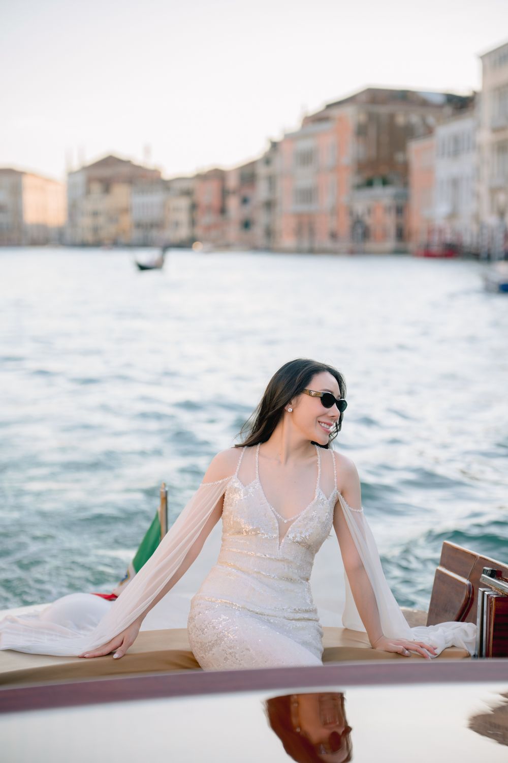 Smiling bride in white lace gown and sunglasses seated on a boat along Venice's Grand Canal.
