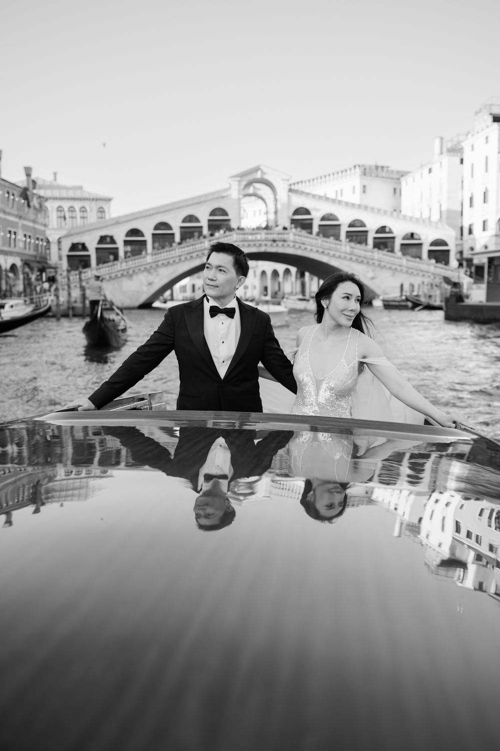 Couple in formal attire poses on a gondola with Venice's Rialto Bridge reflected in black and white.