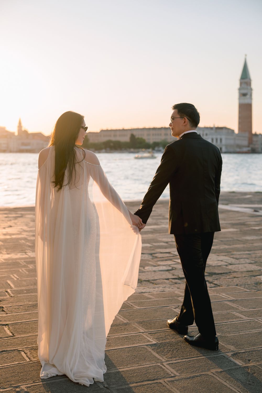 Bride and groom hold hands on Venice waterfront at sunset with St. Mark's Campanile in the background.