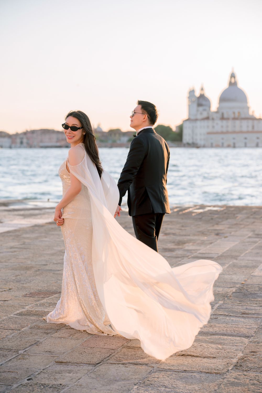 Bride in flowing white gown and groom in black tuxedo pose by Venice waterfront at sunset.